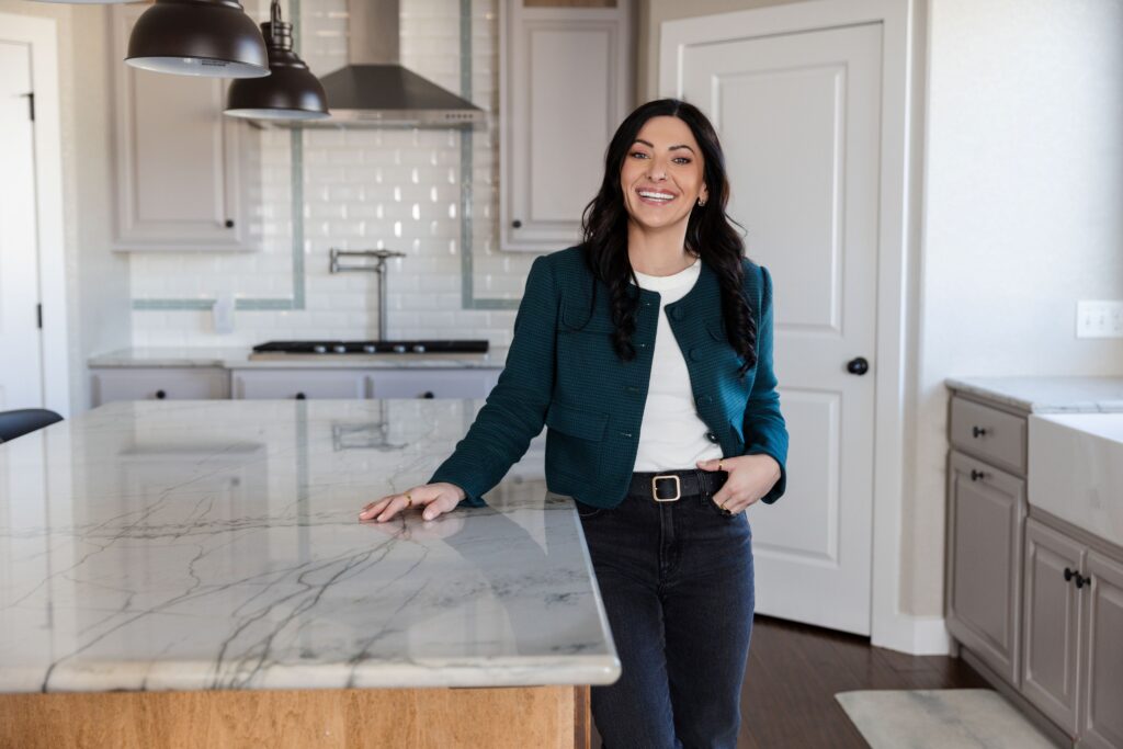 A smiling woman in a teal jacket and dark jeans leans against a marble-topped island in a modern kitchen featuring light grey cabinets, dark pendant lights, and a white subway tile backsplash.