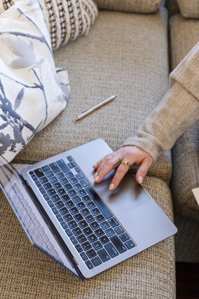A person's hand wearing a gold ring rests on the trackpad of a silver laptop situated on a grey textured sofa, alongside a silver pen and a patterned throw pillow.