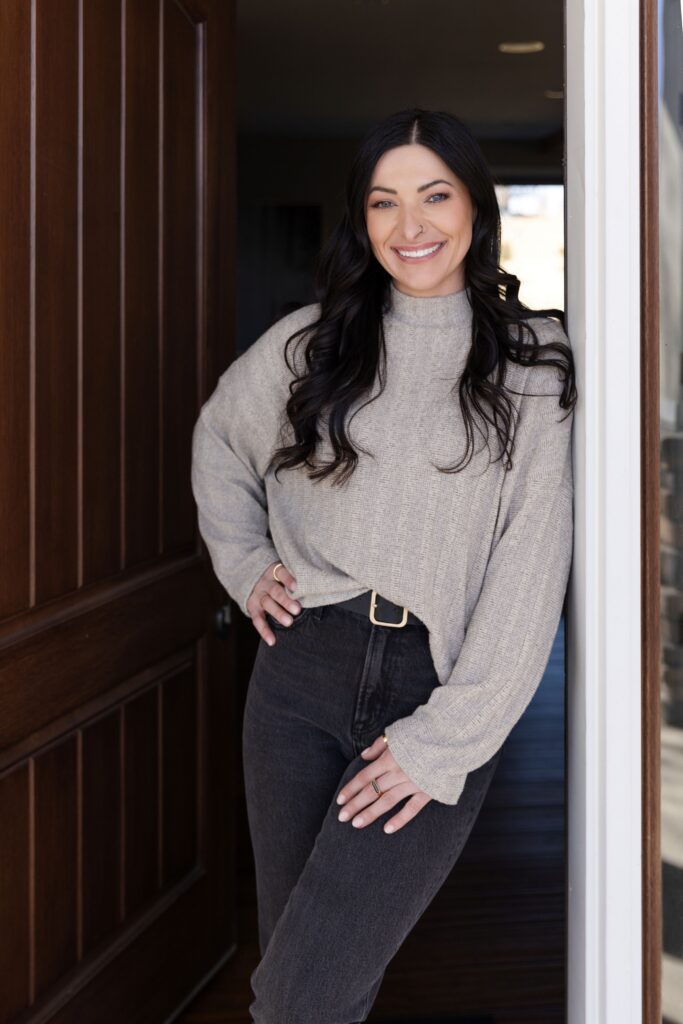 A smiling woman with long dark hair leans against a white doorframe next to an open wooden door, wearing a light grey sweater, dark jeans, and a black belt with a gold buckle.