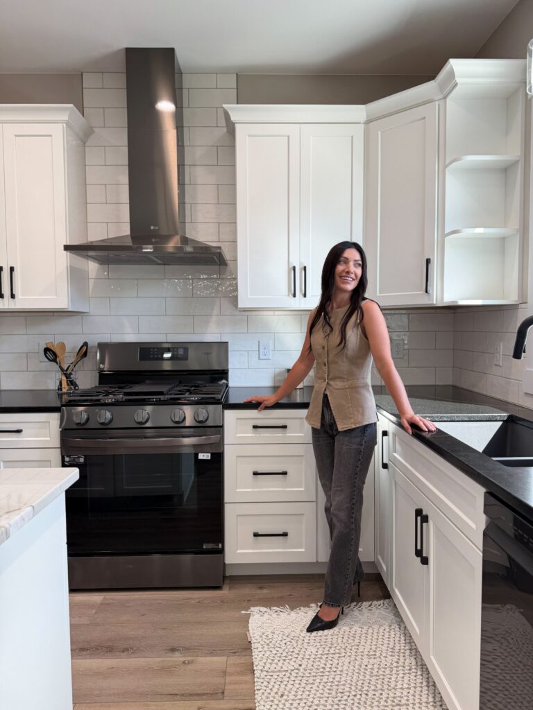 A smiling woman in a beige top and jeans stands in a modern kitchen featuring white cabinets, dark countertops, a subway tile backsplash, and stainless steel appliances.