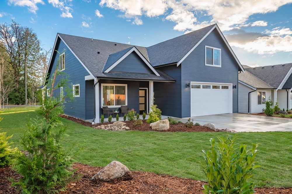 A dark blue house with white trim and a white garage door sits behind a green lawn, landscaped beds with rocks, and a concrete driveway under a partly cloudy sky.